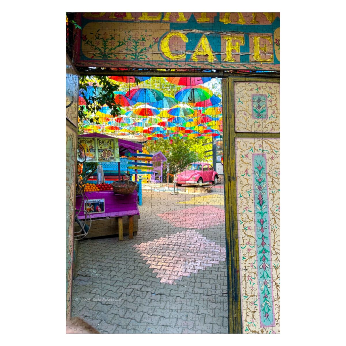 A colorful view through an ornate doorway into a vibrant outdoor street scene with hanging multicolored umbrellas, a small purple cart, a pink vintage car, and a sign reading CAFE at the top.